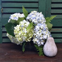 Bouquet of pale blue and white hydrangeas with fern fronds beside a pear-shaped vase