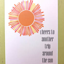 Greeting card with stylized orange sunburst and text 'cheers to another trip around the sun'