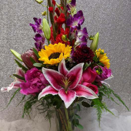Tall mixed bouquet with pink lilies, sunflowers, purple orchids, and red blooms in a clear glass vase