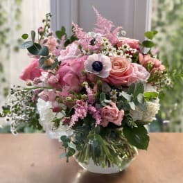 Pink and white mixed bouquet with roses and anemones in a round clear glass vase on a wooden surface.