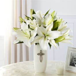 White lilies arranged in a tall white vase with a cross emblem