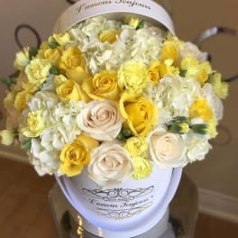 Round hatbox arrangement of yellow and cream roses, carnations, and white hydrangeas in a white container