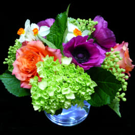 Bright mixed bouquet with orange roses, green hydrangea, purple anemone, and white flowers in a clear glass vase.