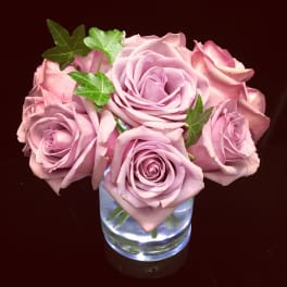 Compact arrangement of mauve-pink roses with ivy in a clear glass cylinder vase.