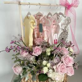 Pink and white rose arrangement in a woven basket with baby dresses behind it