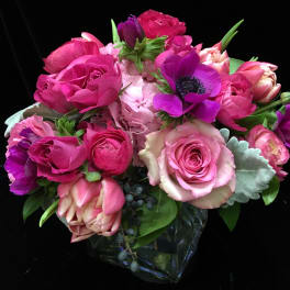 Compact arrangement of pink roses, tulips, and purple anemones in a clear glass cube vase.