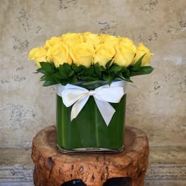 Yellow roses arranged in a square glass vase with a white ribbon