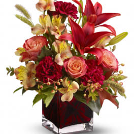 Compact arrangement of red lilies, roses, carnations, and alstroemeria in a square glass vase