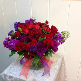 Low round arrangement of red roses and carnations with purple blooms in a clear glass vase with red and purple ribbons.