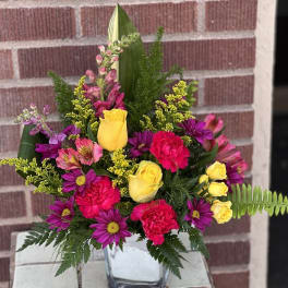 Mixed bouquet of yellow roses, pink carnations, and purple daisies in a glass vase
