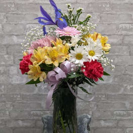 Mixed bouquet of colorful flowers in a tall glass vase with a lavender ribbon