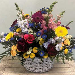 Mixed bouquet in a white wicker basket with bright yellow, pink, blue, and white flowers