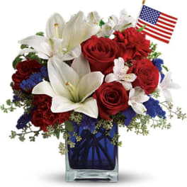 Red, white, and blue flower arrangement with roses and lilies in a blue vase with a small American flag.