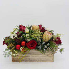 Low red and white flower arrangement with protea and roses in a wooden rectangular box with red ornaments.
