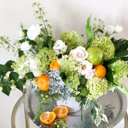 White and green floral arrangement with oranges and a green apple in a white vase