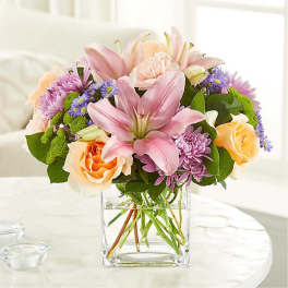 Mixed bouquet of pink lilies, peach roses, and purple flowers in a clear square glass vase
