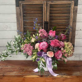 Pink and purple mixed flower arrangement in a white container with ribbon