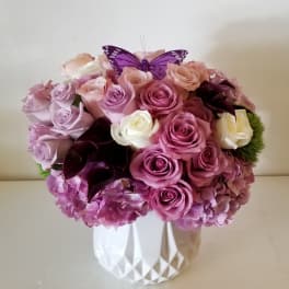 Pink and lavender roses arranged in a white vase with a purple butterfly topper