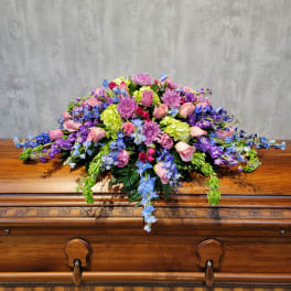 Large floral spray with pink roses, purple blooms, and blue flowers on a wooden casket