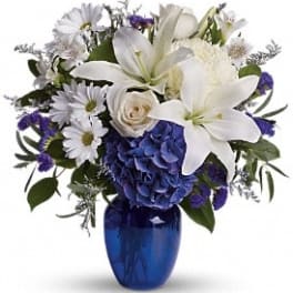 White lilies, daisies, roses and blue hydrangea arranged in a cobalt blue glass vase