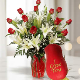 Red roses and white lilies in a glass vase beside a red rose-shaped gift