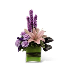 Modern arrangement of pink lily, purple flowers, and dark calla lilies in a green-lined glass cube vase