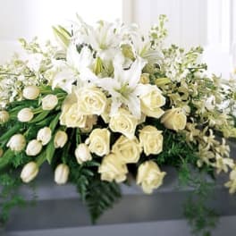 White casket spray of lilies, roses, tulips, and orchids cascading over a gray casket