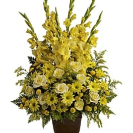Tall yellow sympathy-style arrangement with roses, daisies, and gladiolus in a dark pedestal vase