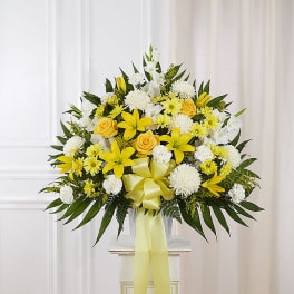 Large yellow and white sympathy arrangement with lilies, roses, and mums in a white urn with a yellow bow