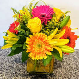 Bright mixed bouquet with gerbera daisies, roses, and lilies in a square vase