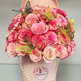 Pink roses arranged in a round pink hat box with butterfly accents and pearl strands.