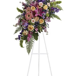 Standing sympathy spray with pink lilies, roses, and mixed purple and yellow flowers on a white easel