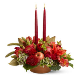 Low centerpiece of red and orange flowers with green hydrangea around two red taper candles in a brown bowl