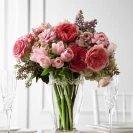 Pink and coral roses, tulips, and mixed blooms arranged in a clear glass vase on a table.
