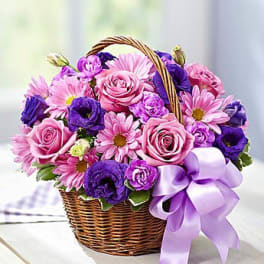 Pink and purple roses, daisies, and blooms arranged in a wicker basket with a lavender bow