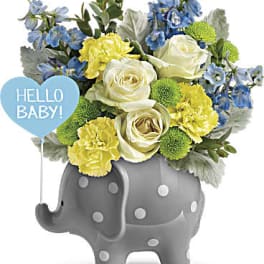 Bouquet of white roses, yellow carnations, and blue flowers in a gray elephant planter with a baby balloon