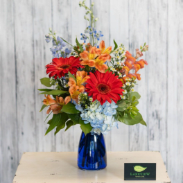 Mixed bouquet of red gerbera daisies, orange blooms, and blue hydrangeas in a blue glass vase