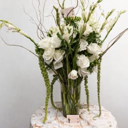 Tall white flower arrangement with roses and trailing green accents in a clear glass vase on a round table.