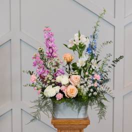 Tall mixed flower arrangement in a white container with pink, peach, white, and blue blooms.