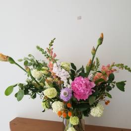 Tall mixed arrangement of pink peony, white hydrangea clusters, snapdragons, and orange accents in a clear glass vase