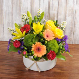 Bright mixed arrangement with yellow lilies and roses, orange gerberas, and red and purple blooms in a white bowl