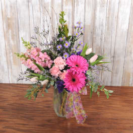 Tall mixed arrangement with pink gerbera daisies, carnations, tulips and purple asters in a glass vase with ribbon