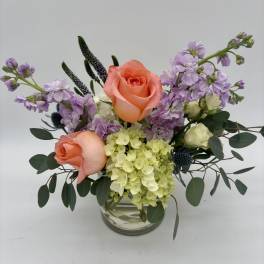 Arrangement of peach roses, green and ivory hydrangeas, and purple blooms in a clear glass vase