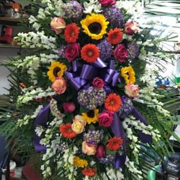 Large standing floral spray with sunflowers, gerbera daisies, roses, and purple ribbon
