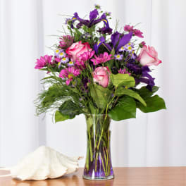 Tall bouquet of pink roses, purple irises, and pink daisies in a clear vase beside a large white seashell.