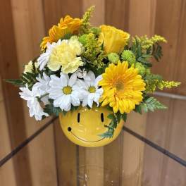 Yellow and white daisies, roses, and carnations in a yellow smiley-face ceramic vase.