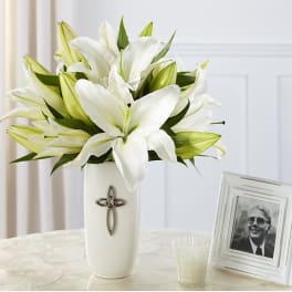 White lilies arranged in a tall white vase beside a framed photo
