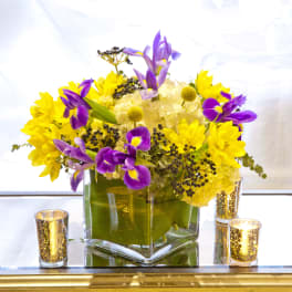 Purple irises and yellow flowers in a square glass vase with candles