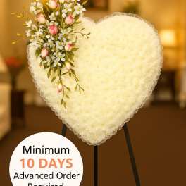 Heart-shaped white sympathy wreath with pink buds and small white flowers on a black easel stand indoors.