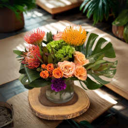 Tropical arrangement with orange protea, roses, and monstera leaves in a glass vase